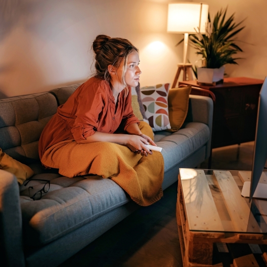 Eine Frau auf der Couch mit einer Fernbedingung in der Hand und gespannten Blick.