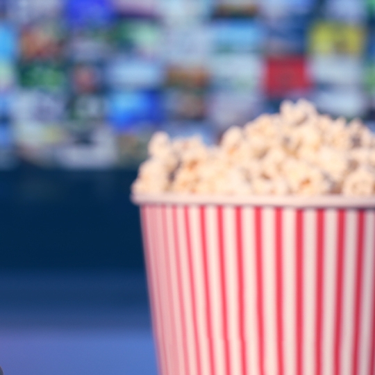 Eine Fernbedienung in der Hand, mit Popcorn und einem Fernseher im Hintergrund.