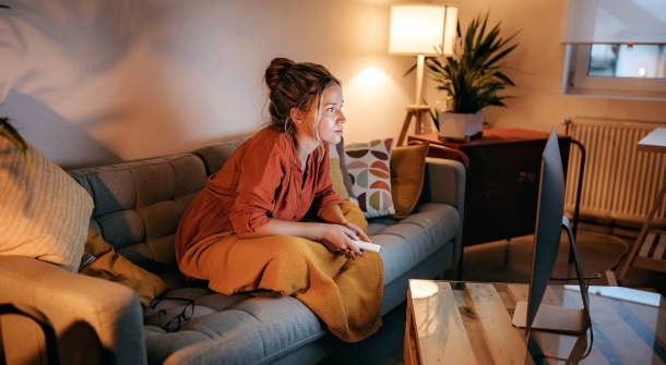 Eine Frau auf der Couch mit einer Fernbedingung in der Hand und gespannten Blick.