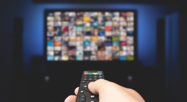 Eine Fernbedienung in der Hand zeigt auf einen Fernseher, auf dem mehrere Sender angezeigt werden.