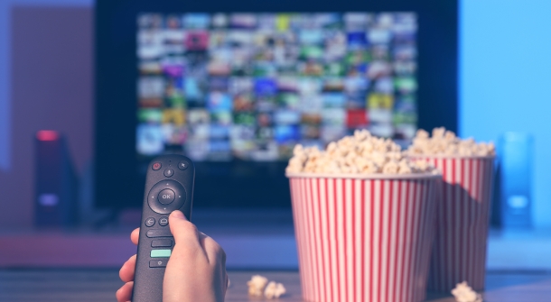Eine Fernbedienung in der Hand, mit Popcorn und einem Fernseher im Hintergrund.